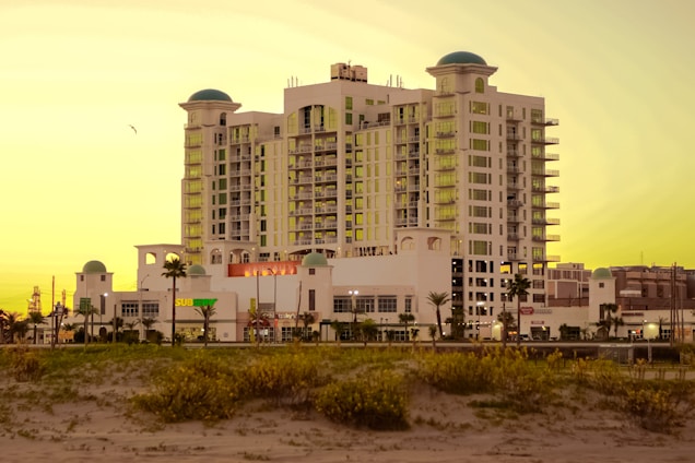 A multi-story hotel building with a white facade and several balconies, featuring rounded green domes on its roof. It is set against a golden sky, likely at sunset, with sandy foreground and green shrubs. Palm trees are visible near the entrance, and there is signage for a Subway restaurant on the ground floor.