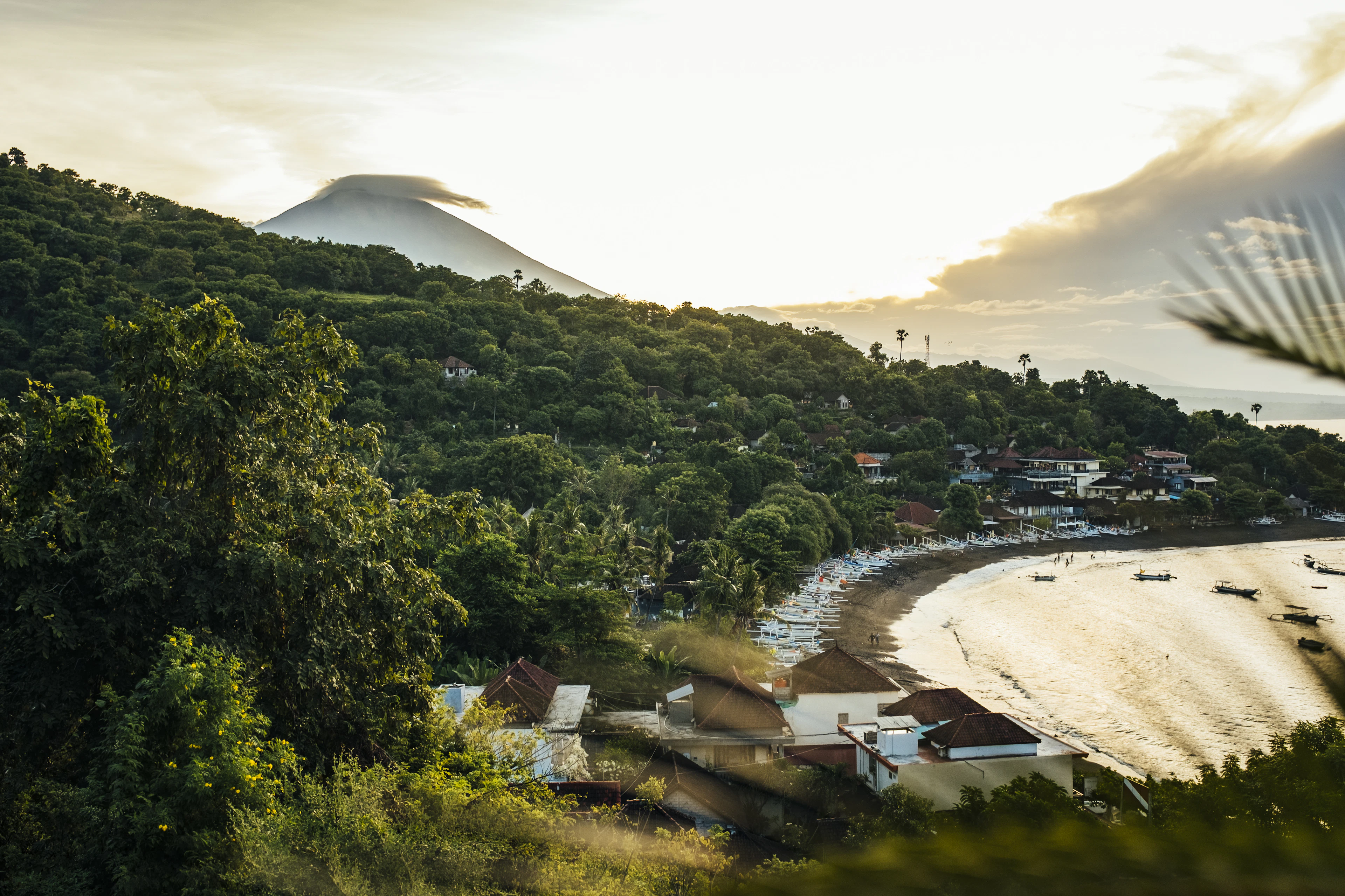 An aerial view at golden hour shows a cloud-wrapped volcano above a green cove village on the coast.