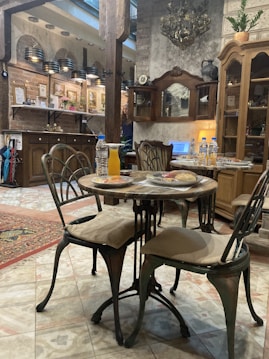 A cozy, rustic-style interior features a wooden dining table with two chairs, set with plates and drinks. The room has vintage decor elements like an ornate chandelier, a wooden cabinet with shelves, and paintings on the walls. Soft lighting adds warmth to the atmosphere.