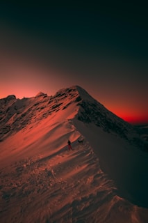 A lone hiker, accompanied by a dog, stands on a snow-covered mountain ridge. The sky is lit with a dramatic gradient of deep red and orange, suggesting either sunrise or sunset. Shadows cast across the snow enhance the rugged features of the mountain landscape.