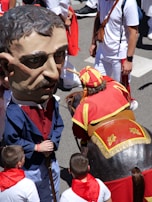 People are participating in a festive event with one person wearing an oversized mask and traditional attire. Another person is dressed in a costume representing a horse. The crowd is dressed in white with red scarves, typical of celebrations like the Running of the Bulls.
