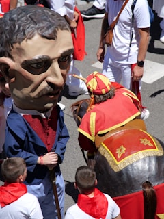 People are participating in a festive event with one person wearing an oversized mask and traditional attire. Another person is dressed in a costume representing a horse. The crowd is dressed in white with red scarves, typical of celebrations like the Running of the Bulls.