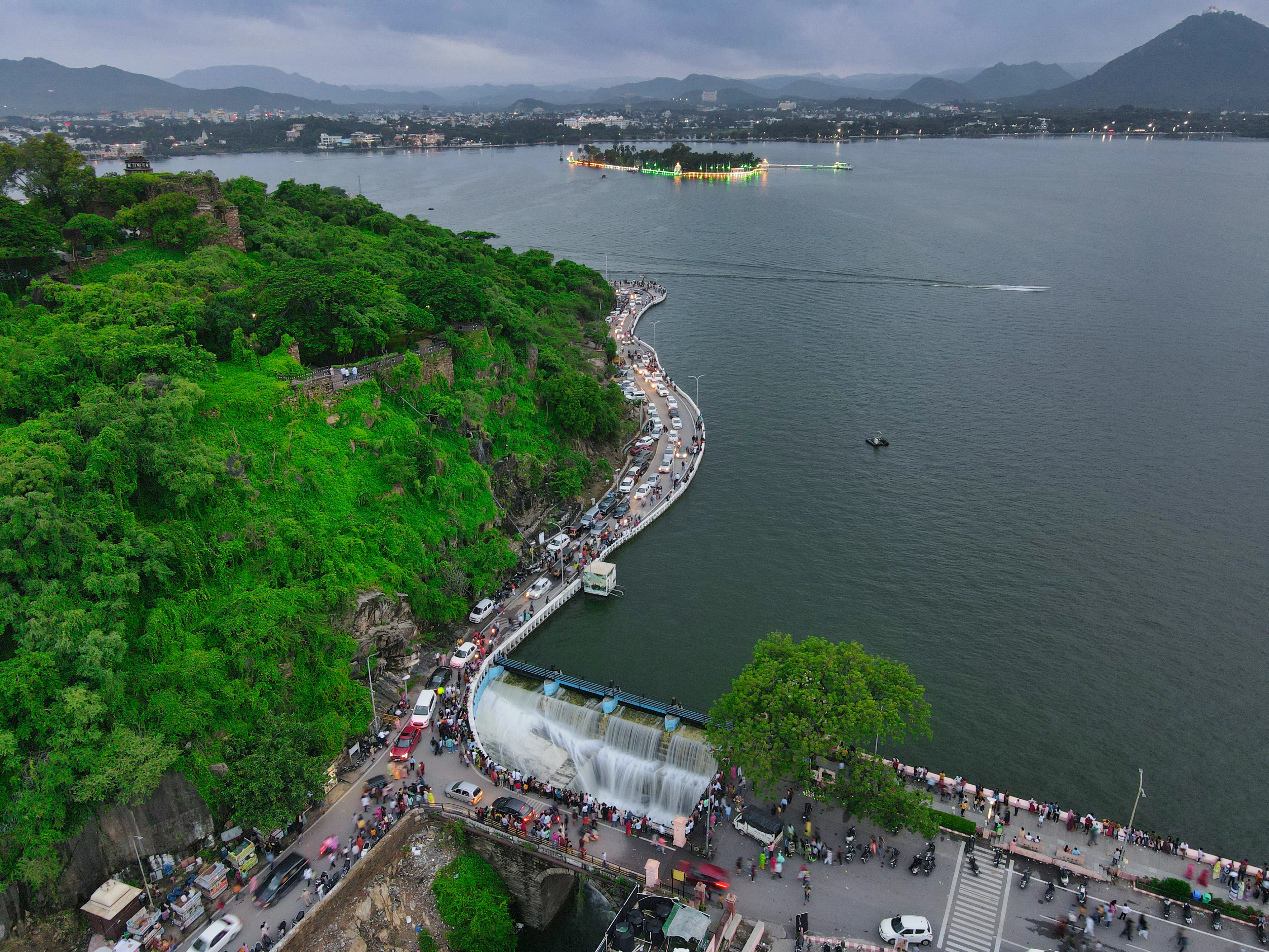 Fateh Sagar Lake in Udaipur brimming over its spillway after monsoon rain, a white curtain of water pouring beside the causeway while crowds line the lakefront promenade, jungle-green hills and the lit island of Nehru Park beyond