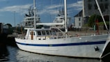 A white yacht with a blue stripe is docked at a harbor. In the background, two larger ships and a stone building are visible, alongside a harbor area with walkways and some greenery.