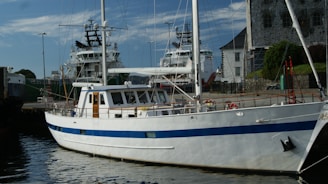 A white yacht with a blue stripe is docked at a harbor. In the background, two larger ships and a stone building are visible, alongside a harbor area with walkways and some greenery.