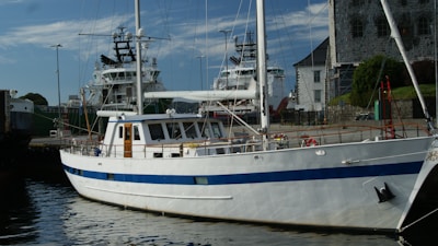 A white yacht with a blue stripe is docked at a harbor. In the background, two larger ships and a stone building are visible, alongside a harbor area with walkways and some greenery.
