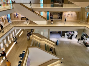 A modern, multi-level indoor space with wooden flooring and a large open area. The levels are connected by wide staircases, and various people are seen walking, sitting, and interacting throughout. There are displays and retail sections with items and people browsing.