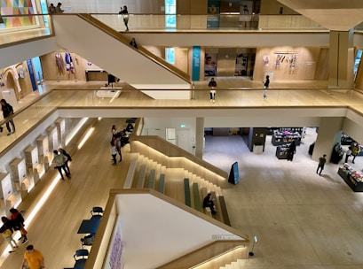 A modern, multi-level indoor space with wooden flooring and a large open area. The levels are connected by wide staircases, and various people are seen walking, sitting, and interacting throughout. There are displays and retail sections with items and people browsing.