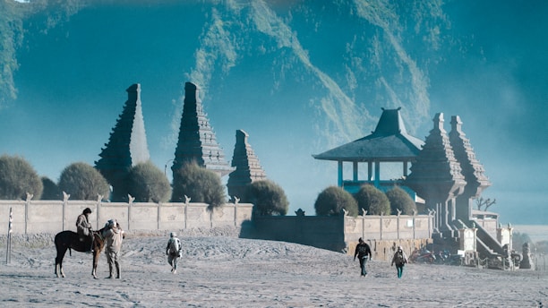 A majestic ancient temple structure is surrounded by trees and misty hills in the background. Several people are seen walking near the temple, with one person riding a horse and another standing beside it.