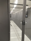 A corridor inside a data center with rows of server racks enclosed within glass panels. Overhead lighting brightly illuminates the area, and perforated floors suggest climate control measures.