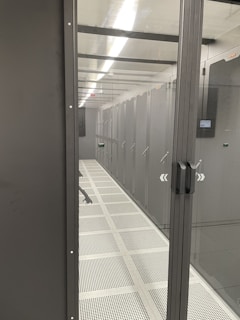 A corridor inside a data center with rows of server racks enclosed within glass panels. Overhead lighting brightly illuminates the area, and perforated floors suggest climate control measures.