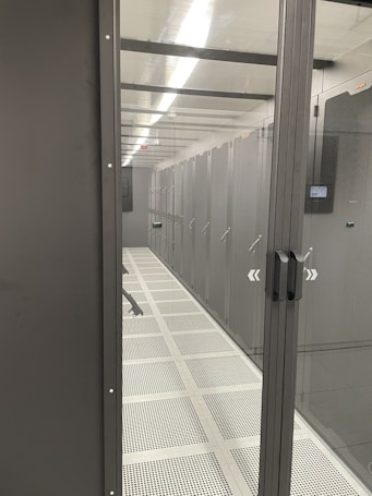 A corridor inside a data center with rows of server racks enclosed within glass panels. Overhead lighting brightly illuminates the area, and perforated floors suggest climate control measures.