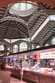 A spacious indoor market hall with high, ornate ceilings featuring intricate architectural details and large arched windows. Several market stalls display seafood with vendors attending to customers. Bright lighting highlights the fresh produce, and signage is visible above each stall.