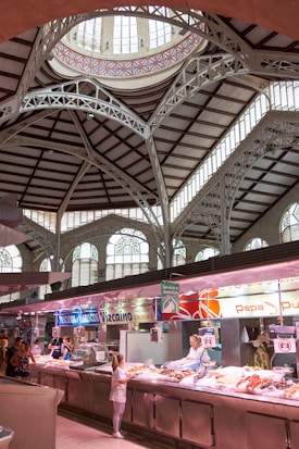 A spacious indoor market hall with high, ornate ceilings featuring intricate architectural details and large arched windows. Several market stalls display seafood with vendors attending to customers. Bright lighting highlights the fresh produce, and signage is visible above each stall.