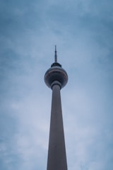 A tall telecommunications tower rises into a cloudy sky, with a spherical observation deck and an antenna at the top.