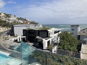 A coastal residential area with modern architecture overlooking the ocean. The scene includes a luxury building labeled 'AURUM', surrounded by well-maintained natural greenery. A road curves along the hillside with several parked cars and more houses extending into the distance. The sky is partly cloudy, blending with the blue of the ocean.