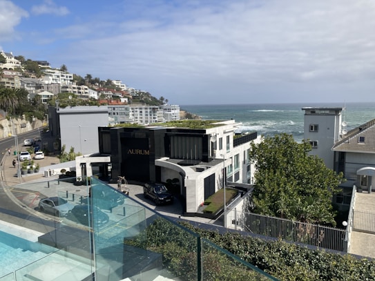 A coastal residential area with modern architecture overlooking the ocean. The scene includes a luxury building labeled 'AURUM', surrounded by well-maintained natural greenery. A road curves along the hillside with several parked cars and more houses extending into the distance. The sky is partly cloudy, blending with the blue of the ocean.