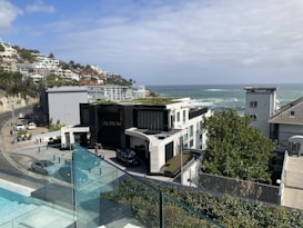 A coastal residential area with modern architecture overlooking the ocean. The scene includes a luxury building labeled 'AURUM', surrounded by well-maintained natural greenery. A road curves along the hillside with several parked cars and more houses extending into the distance. The sky is partly cloudy, blending with the blue of the ocean.