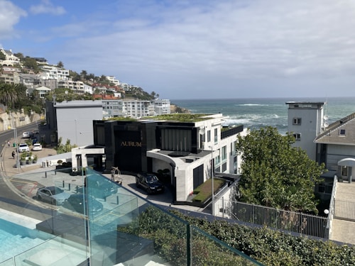 A coastal residential area with modern architecture overlooking the ocean. The scene includes a luxury building labeled 'AURUM', surrounded by well-maintained natural greenery. A road curves along the hillside with several parked cars and more houses extending into the distance. The sky is partly cloudy, blending with the blue of the ocean.