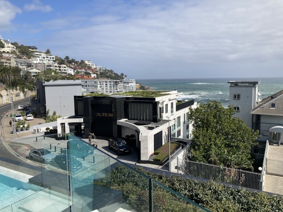 A coastal residential area with modern architecture overlooking the ocean. The scene includes a luxury building labeled 'AURUM', surrounded by well-maintained natural greenery. A road curves along the hillside with several parked cars and more houses extending into the distance. The sky is partly cloudy, blending with the blue of the ocean.