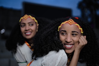 Two smiling women with curly hair and decorative headbands stand closely together. They wear traditional clothing with vibrant patterns. The setting appears to be outdoors with dim lighting, suggesting evening or nighttime.