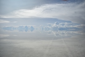 A vast and reflective salt flat mirrors the expansive sky above. Puffy clouds are scattered across the horizon, blending seamlessly into their reflections on the ground, creating an illusion of endless space.