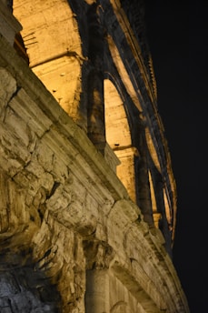 A close-up view of an ancient stone structure illuminated warmly against a dark sky. The photo captures arches and the textured stonework typically found in historical Roman architecture.