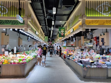 A bustling market scene with shoppers examining a variety of fresh produce and assorted goods displayed on long stalls. The market is well-lit with overhead lighting, and the decor includes a bright color scheme with signs and banners featuring graphics and writings.
