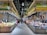 A bustling market scene with shoppers examining a variety of fresh produce and assorted goods displayed on long stalls. The market is well-lit with overhead lighting, and the decor includes a bright color scheme with signs and banners featuring graphics and writings.
