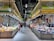 A bustling market scene with shoppers examining a variety of fresh produce and assorted goods displayed on long stalls. The market is well-lit with overhead lighting, and the decor includes a bright color scheme with signs and banners featuring graphics and writings.