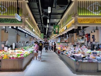 A bustling market scene with shoppers examining a variety of fresh produce and assorted goods displayed on long stalls. The market is well-lit with overhead lighting, and the decor includes a bright color scheme with signs and banners featuring graphics and writings.