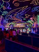 A vibrant and colorful space filled with neon light designs on the ceiling and walls, creating an immersive atmosphere. People are gathered in a queue, likely waiting to enter an attraction. The environment is reminiscent of an aquarium or theme park setting, with signs and decorations enhancing the lively mood.