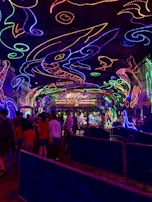 A vibrant and colorful space filled with neon light designs on the ceiling and walls, creating an immersive atmosphere. People are gathered in a queue, likely waiting to enter an attraction. The environment is reminiscent of an aquarium or theme park setting, with signs and decorations enhancing the lively mood.