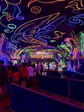 A vibrant and colorful space filled with neon light designs on the ceiling and walls, creating an immersive atmosphere. People are gathered in a queue, likely waiting to enter an attraction. The environment is reminiscent of an aquarium or theme park setting, with signs and decorations enhancing the lively mood.