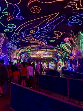 A vibrant and colorful space filled with neon light designs on the ceiling and walls, creating an immersive atmosphere. People are gathered in a queue, likely waiting to enter an attraction. The environment is reminiscent of an aquarium or theme park setting, with signs and decorations enhancing the lively mood.