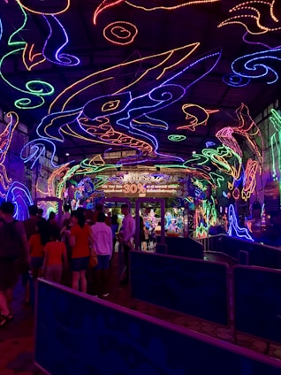 A vibrant and colorful space filled with neon light designs on the ceiling and walls, creating an immersive atmosphere. People are gathered in a queue, likely waiting to enter an attraction. The environment is reminiscent of an aquarium or theme park setting, with signs and decorations enhancing the lively mood.