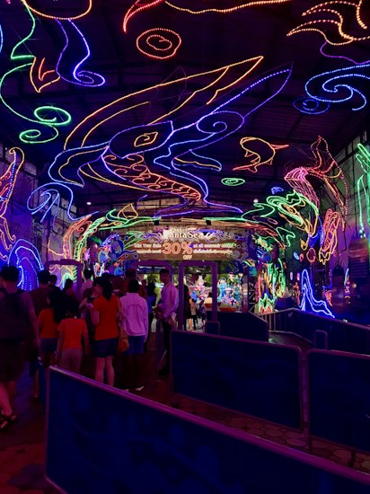 A vibrant and colorful space filled with neon light designs on the ceiling and walls, creating an immersive atmosphere. People are gathered in a queue, likely waiting to enter an attraction. The environment is reminiscent of an aquarium or theme park setting, with signs and decorations enhancing the lively mood.
