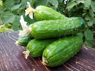 Five fresh green cucumbers with small white spots are stacked on each other on a wooden table outdoors, with green foliage in the background.