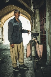 An elderly man wearing traditional clothing stands next to a worn bicycle. The setting is an old, narrow street with an arched pathway. The man is wearing a dark jacket and cap, and the bicycle has fabric bags over its back wheel.