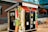 A colorfully painted kiosk labeled as a community fridge is situated outdoors. The structure is adorned with vibrant illustrations of vegetables such as corn, peppers, and peas. It has a welcoming message, inviting people to 'Take what you need, leave what you can.' The kiosk is positioned in front of a brick building, and there are bicycles parked nearby. Additionally, a hanging basket of flowers and a wooden bench are present in the vicinity.