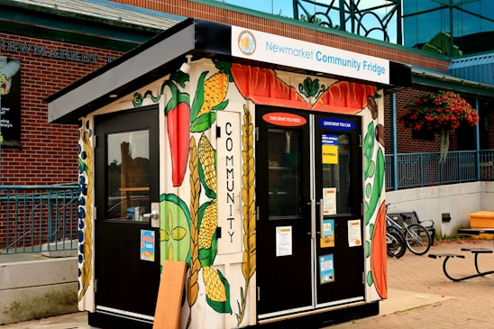 A colorfully painted kiosk labeled as a community fridge is situated outdoors. The structure is adorned with vibrant illustrations of vegetables such as corn, peppers, and peas. It has a welcoming message, inviting people to 'Take what you need, leave what you can.' The kiosk is positioned in front of a brick building, and there are bicycles parked nearby. Additionally, a hanging basket of flowers and a wooden bench are present in the vicinity.