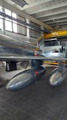 A metallic aircraft featuring a distinctive design with two drop tanks under the wings is inside a hangar. The aircraft showcases a polished, reflective surface with markings. The hangar has a high ceiling with large windows allowing natural light to illuminate the space.