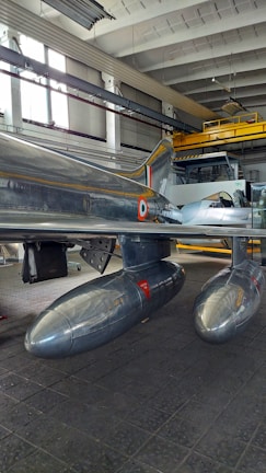 A metallic aircraft featuring a distinctive design with two drop tanks under the wings is inside a hangar. The aircraft showcases a polished, reflective surface with markings. The hangar has a high ceiling with large windows allowing natural light to illuminate the space.