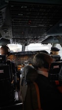 The cockpit of an airplane with two pilots seated and wearing headsets. The cockpit is filled with numerous controls, switches, and displays, indicating a commercial aircraft. The lighting inside is subdued, with sunlight visible through the windows.