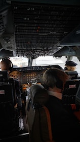 The cockpit of an airplane with two pilots seated and wearing headsets. The cockpit is filled with numerous controls, switches, and displays, indicating a commercial aircraft. The lighting inside is subdued, with sunlight visible through the windows.