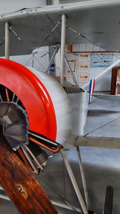 An old biplane with a visible wooden propeller and a radial engine. The plane's body is mainly white with a red nose cone and blue and red stripes on the tail. The background includes display panels with various aircraft illustrations.
