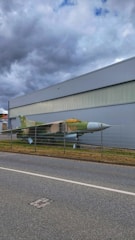 A military jet is displayed behind a metal fence, positioned along a road. The aircraft has a green and brown camouflage paint scheme. It is situated next to a large, industrial-looking building under a cloudy sky.