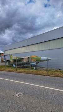 A military jet is displayed behind a metal fence, positioned along a road. The aircraft has a green and brown camouflage paint scheme. It is situated next to a large, industrial-looking building under a cloudy sky.