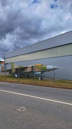 A military jet is displayed behind a metal fence, positioned along a road. The aircraft has a green and brown camouflage paint scheme. It is situated next to a large, industrial-looking building under a cloudy sky.