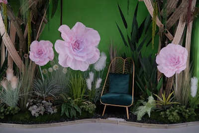 A decorative setting featuring large pink paper flowers against a vibrant green background, with various lush green plants and a stylish chair with a dark green cushion. The scene includes wooden elements and pampas grass, creating a blend of natural and artistic textures.